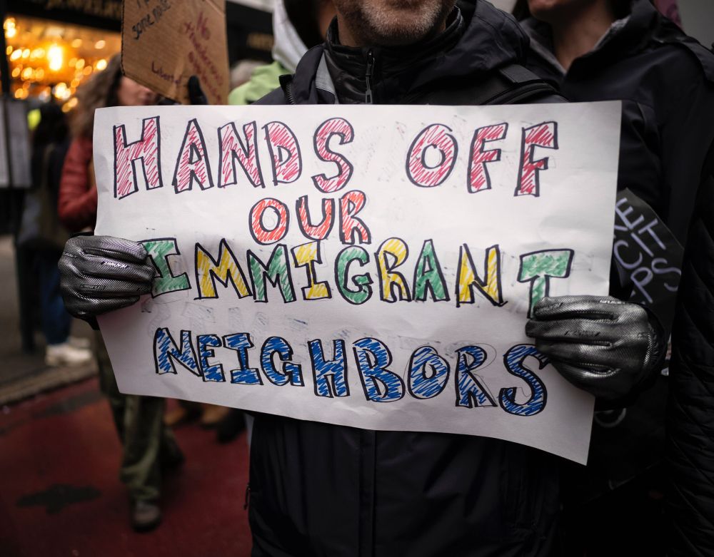 A person holds a handmade protest sign that reads "hands off our immigrant neighbors" amid a rally.