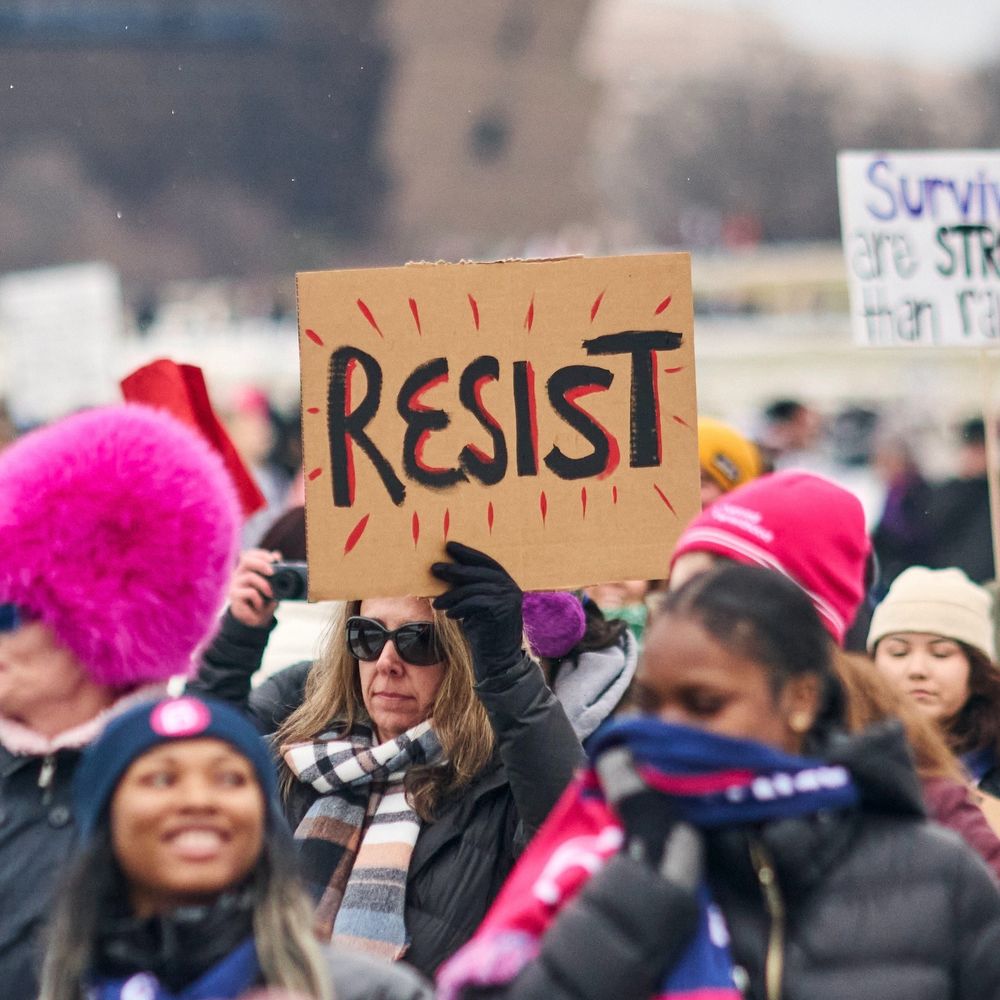Image of a woman at a protest holding a cardboard signing reading "RESIST".