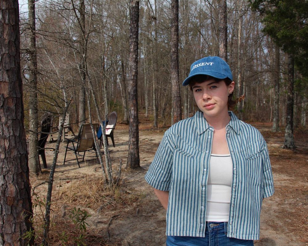 Image of a person in the woods with their hands behind their backs wearing a blue denim dissent hat.