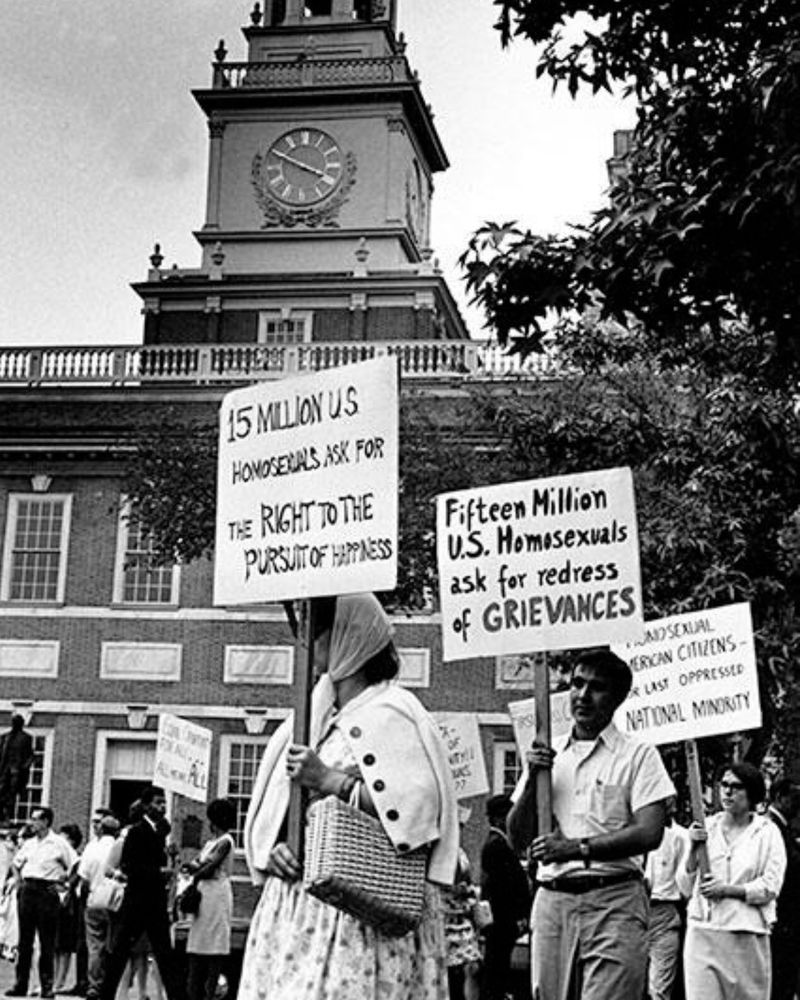 A black and white photo from the 1950s of people protesting outside a courthouse. Picket signs are shown reading "15 million U.S. homosexuals ask for the right to the pursuit of happiness" and "fifteen million U.S. homosexuals ask for a redress of grievances"