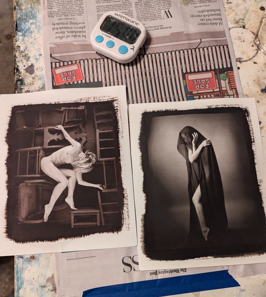 Two kallitype on a table drying from printing. The left is a photo of model Vivian Cove posing nude on a number of chairs at all angles (a gold-toned kallitype - black and white with slight red-purple tones) and the right photo is model Zoe West, standing nude but covered with sheer black fabric with a general dark tone to it (platinum-palladium toned - has some yellow-brown tones to it). Captured also is my timer for timing washing of prints.