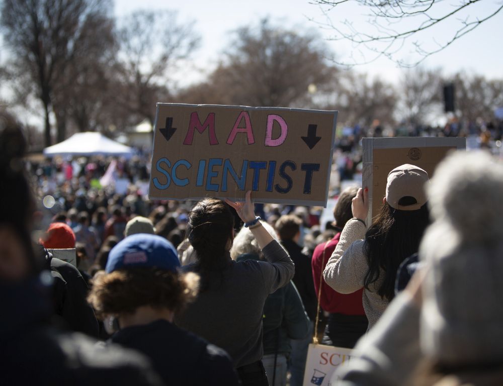 Person in the crowd holding a sign reading MAD SCIENTIST (pointing to herself).