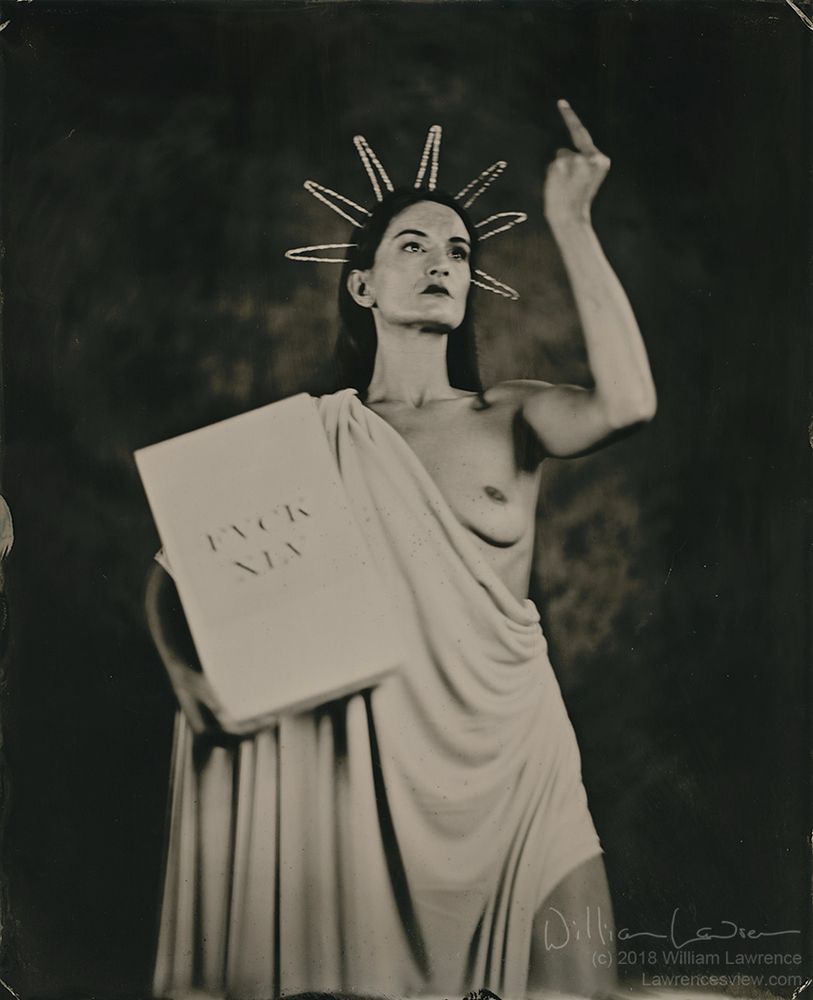A model poses as Lady Liberty standing draped, with one breast exposed and a spiked headdress. She caries a tablet saying 