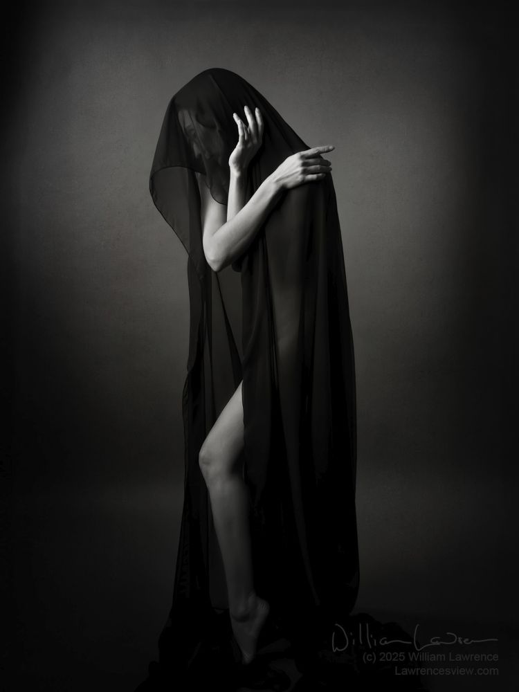 Black and white photograph, slightly toned, of model Zoe C West, posing nude but covered by a draping of sheer black fabric. Only her arms, looking like arms of a clock, and one leg is clearly visible in this low key image.