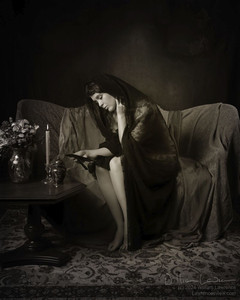 Model Jessa Ray Muse poses sitting on a draped settee, herself draped in sheer black fabric.  She gazes sadly at a photography, with an urn, a melting candle, and a vase of flowers off to the side.