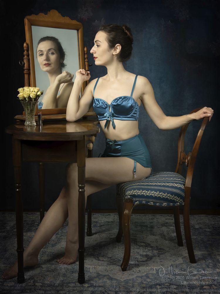 Model Zoe West poses seated in front of a dressing table, wearing a turquoise vintage looking bra and panty set. She gazes in the mirror, and her reflection in the mirror engages the viewer.