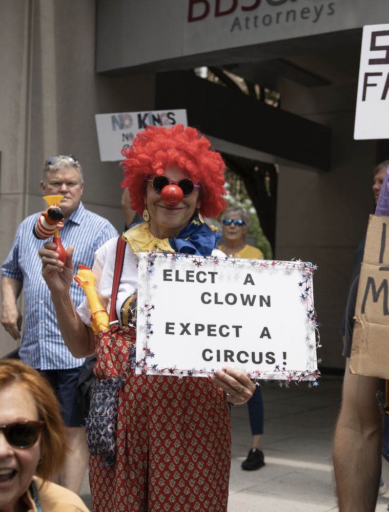 Woman dressed as clown with red wig and red nose, holding sign saying 