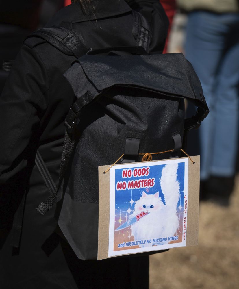 A sign on a person's backpack with an illustrated cat with a knife in it's mouth reads NO GODS / NO MASTERS / AND ABSOLUTELY NO FUCKING KINGS!