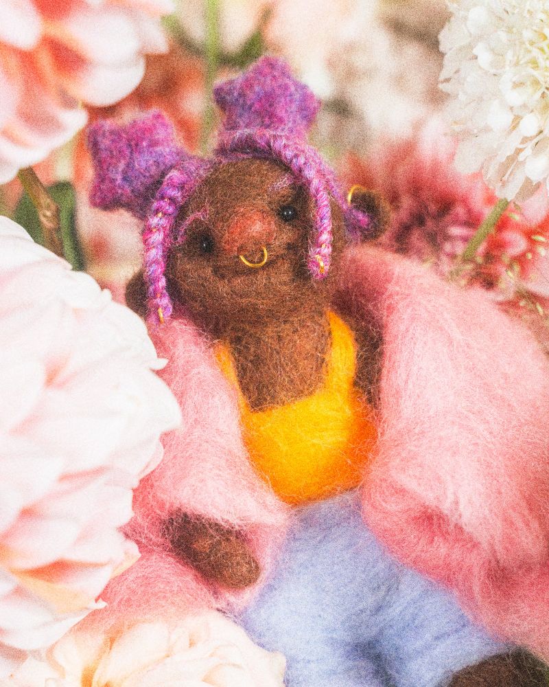 needle felted brown doll, with pink hair shaped into star buns and braids, a  gold septum ring adorning their round pink nose. they are wearing a fuzzy pink coat, a yellow tank top, and blue shorts. they are surrounded by pink and cream colored dahlias and peonies in front of a green backdrop. 