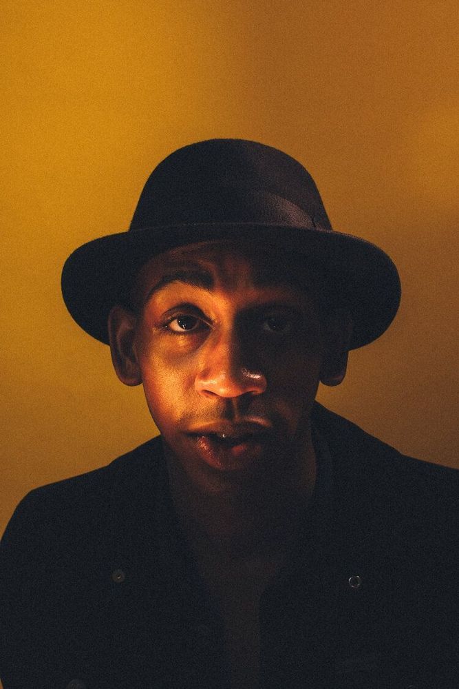 Photograph of a Black man in a black fedora and dark jacket, standing in front of a dappled golden backdrop and looking directly into the lens. Half of his face is illuminated by dramatic and warm lighting, while the rest is shrouded in shadow.
