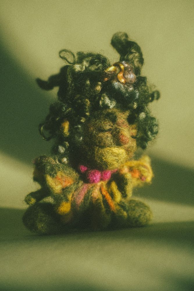 warm and green photograph of a little felted creature in the same color palette. his eyes are closed, his nose is a little red. his cape is adorned with pink pompoms. his head is crowned with disheveled green locks. 