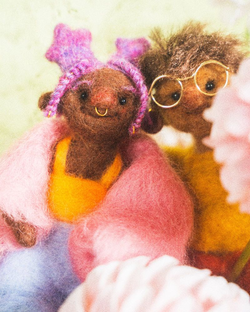 needle felted doll, with pink hair shaped into star buns and braids, a gold septum ring adorning their round pink nose. they are wearing a fuzzy pink coat, a yellow tank top, and blue shorts. next to them is another doll with short brown hair and round golden glasses, clad in a mustard shirt and orange pants. his forehead close to theirs. they are surrounded by pink and cream colored dahlias and peonies in front of a green backdrop. 