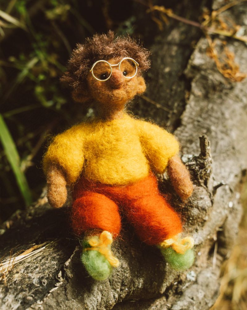 a warm photograph of a little felted guy, he's got light brown skin tone, tiny beaded eyes surrounded by large gold metal glasses. he's wearing a fuzzy yellow top, tiny green shoes with yellow shoe laces, and orange pants. he's sitting on a tree branch, surrounded by foliage.