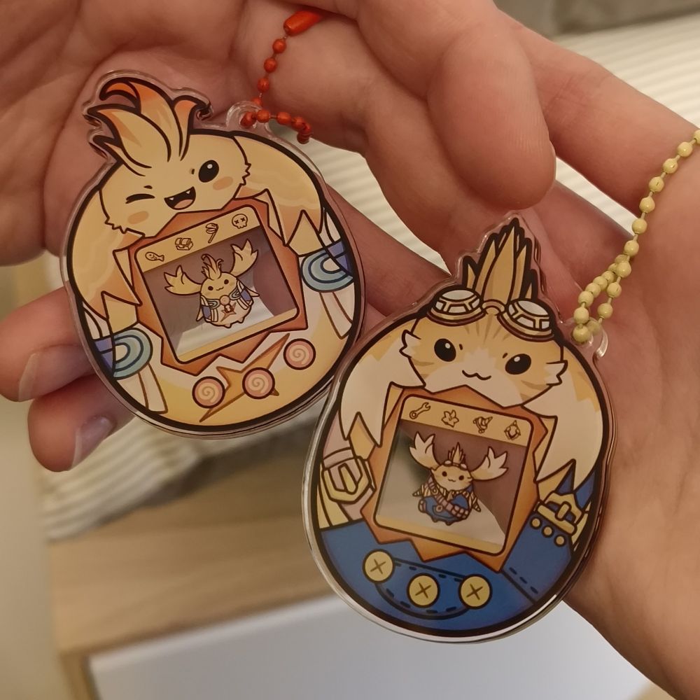 Se ven dos manos sujetando un llavero estilo Tamagotchi cada una. Son personajes de la saga Xenoblade. El de la izquierda es Riki; el de la derecha, Tora.