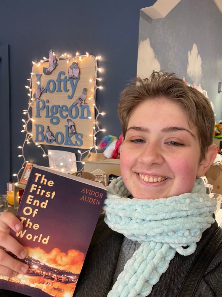 A picture of me, a white butch person in a black coat and blue scarf, holding a book titled "The First End of the World". Behind me is a sign that says "Lofty Pigeon Books".