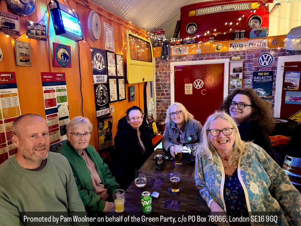 6 people around a pub table with a lot of decorations on the wall. Caption at the bottom reads: Promoted by Pam Woolner on behalf of the Green Party, c/o PO Box 78066, London SE16 9GQ