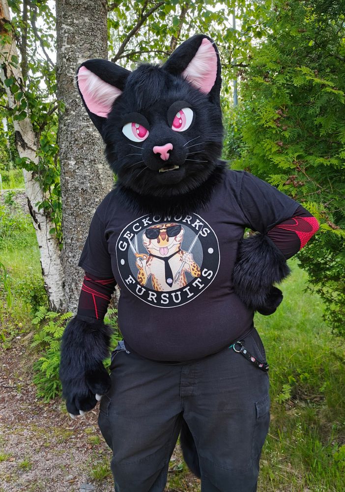 Black cat partial fursuiter. The cat has magenta pink eyes and light pink ears & nose, and is wearing a Geckoworks t-shirt.