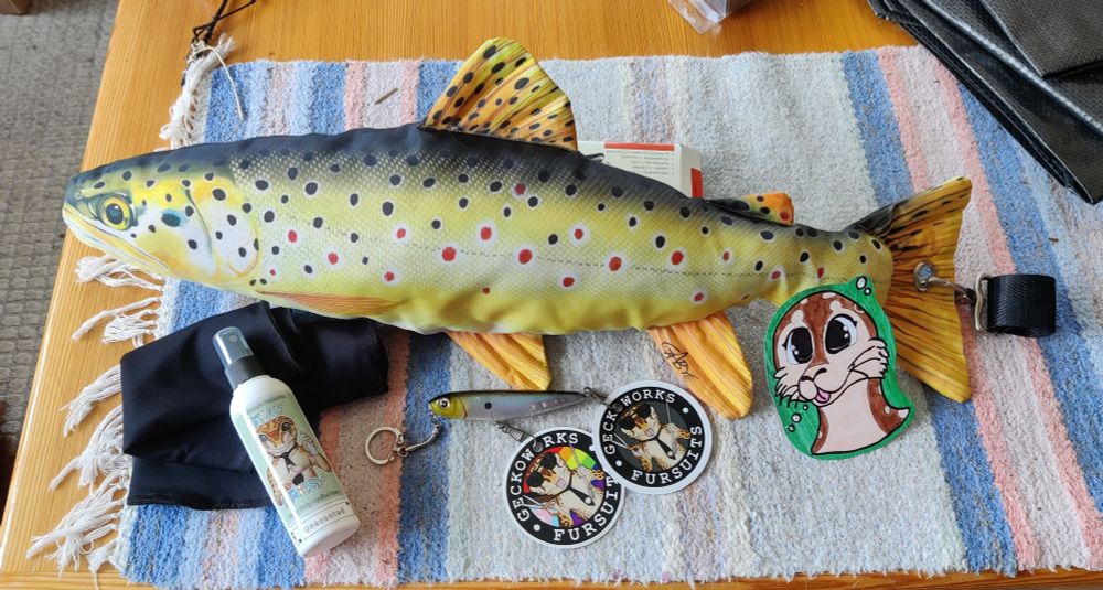Seal fursuit extras kit: 60cm realistic fish (brown trout) plush with clip-to-belt attachment on tail, fish-themed badge/charm holder, traditional headshot badge by Mongrel with custom name & lamination + standard care kit with fursuit spray, balaclava & brush. 