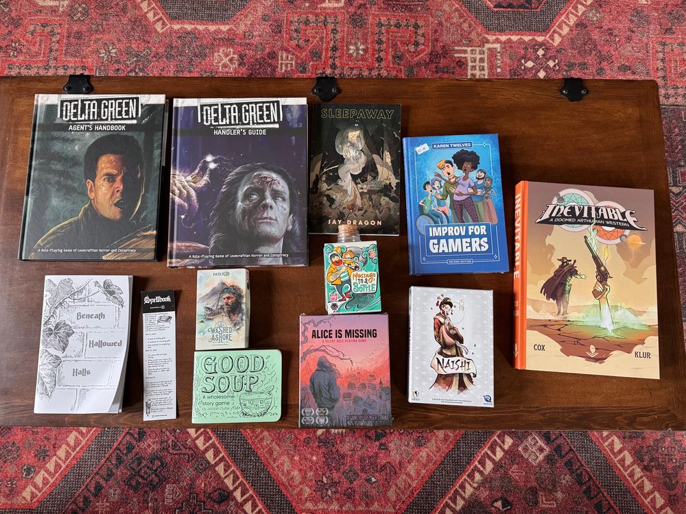 Jess’s GenCon haul is arranged on a wooden coffee table sitting on a red rug. The games on the table are the Delta Green Agent and Handler Handbooks, Sleepaway by Jay Dragon, Improv for Gamers by Karen Twelves, Inevitable by Zachary Cox, Naishi, Message  in a Bottle, Alice is Missing, Good Soup by Jordan Dube, Spellbook by Conlin Durbin, Washed Ashore by Johannes Krenner, and Beneath Hallowed Halls by Ali Mark & Nell Quinn-Gibney. 