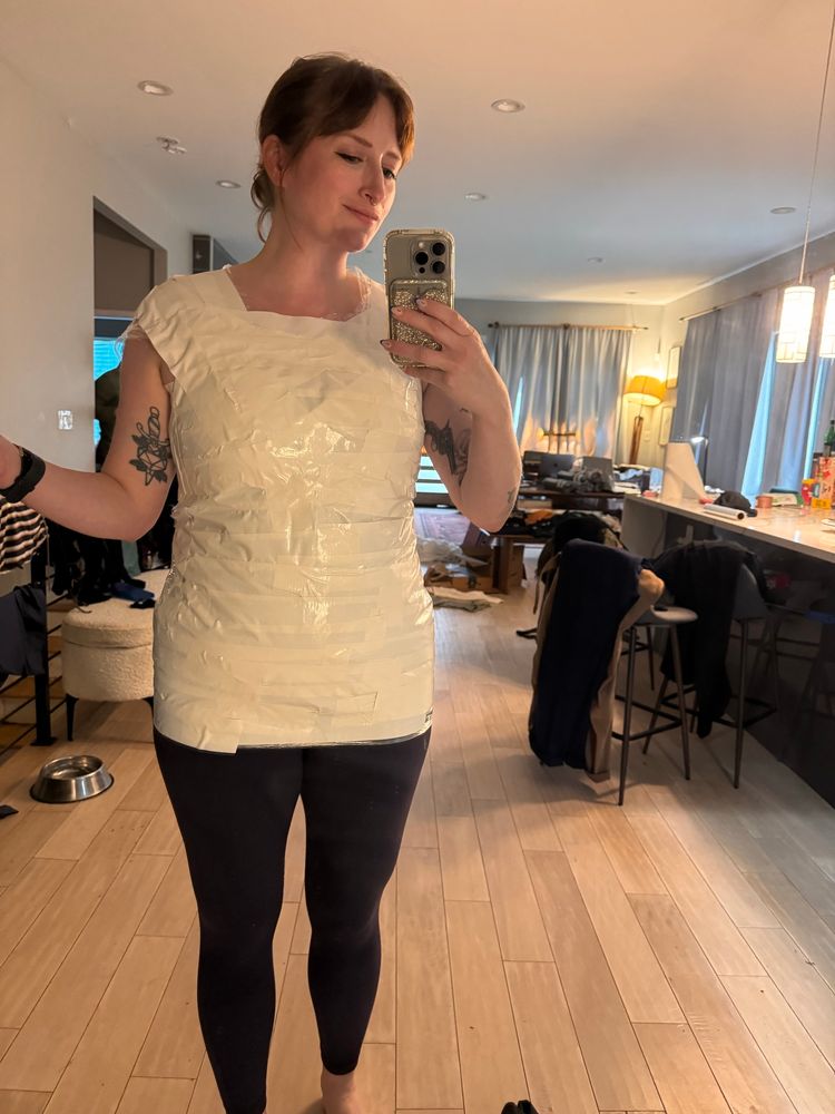 Jess, a white woman with red hair, stands in her (messy) kitchen, wrapped up in Saran Wrap and duct tape to make an armor pattern. 