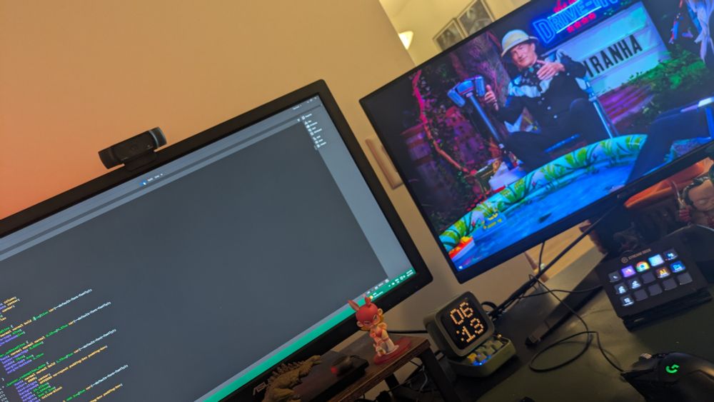 Two computer monitors showing The Last Drive In and a bunch of CSS code. Small Godzilla and other toys are littered on the desk.