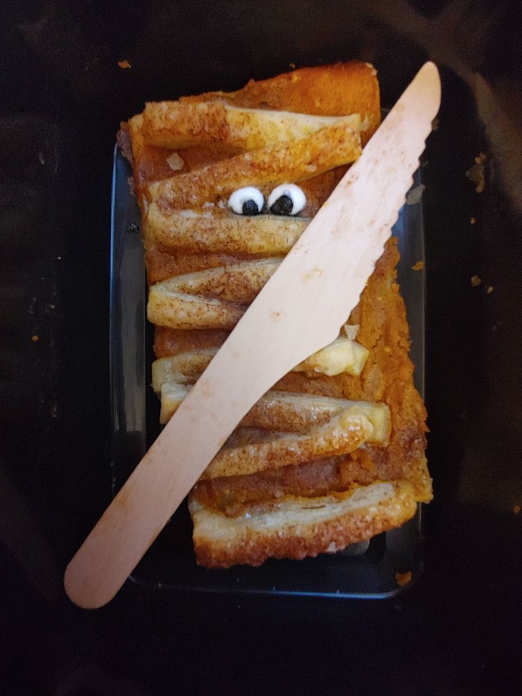 Mummy shaped pumpkin pastry in a plastic box appears to be holding a wooden knives that landed on it