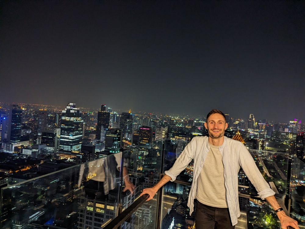 A figure (Chris Park-Kennaby) smiling at the camera in the foreground with the Bangkok skyline in the background