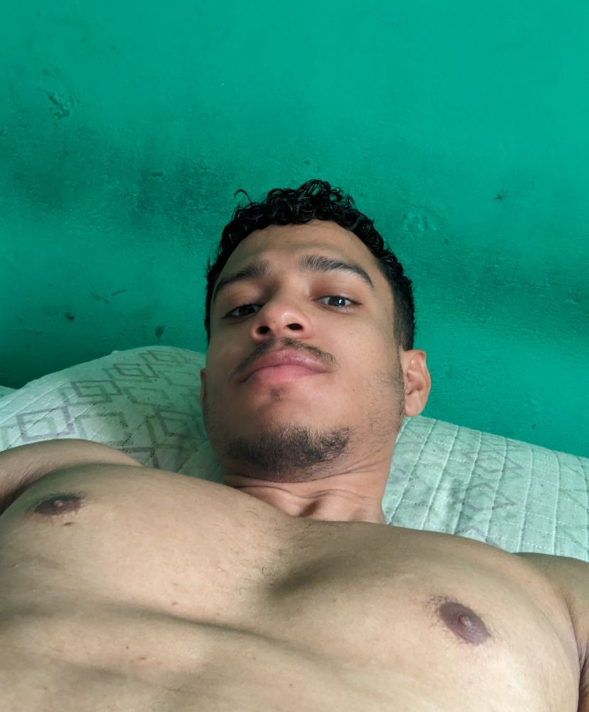 Homem pardo com cabelos cacheados pretos curto, olhos castanhos, tirando uma selfie no ângulo de baixo pra cima, destacando os peitos enquanto está deitado na cama e aí fundo, uma parede verde.