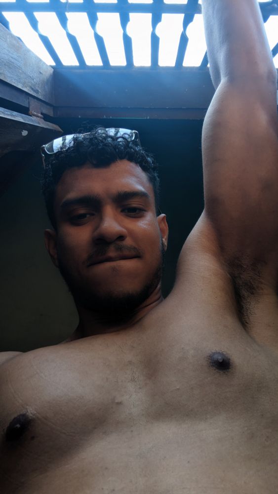 Selfie no ângulo de baixo pra cima, mostrando homem pardo, com cabelos cacheados pretos, usando óculos de grau retangular sobre a cabeça, barba por fazer e mostrando o suvaco direito por estar com o braço segurando as grandes da janela, ressaltando o bíceps e o peitoral.