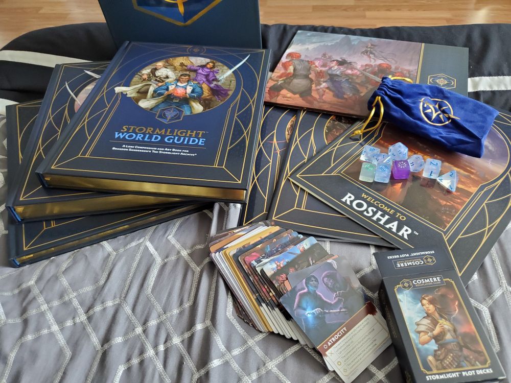 A collection of books, handouts, dice, and cards for a role-playing game. Some of the text reads Stormlight World Guide and Welcome to Roshar. They sit atop grey and black bedding.