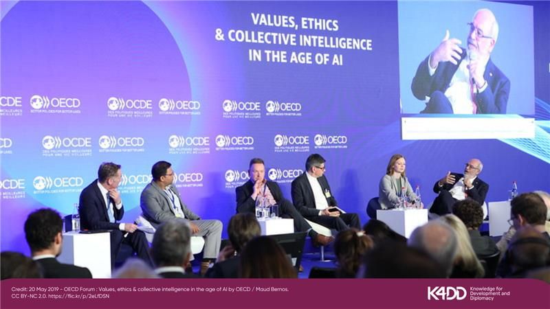 A photo of a group of people smartly dressed sitting as a panel on a stage. There is a large blue background behind them with text that reads 'Values, Ethics, and Collective Intelligence in the Age of AI', along with the OECD logo. There is an audience watching them speak.