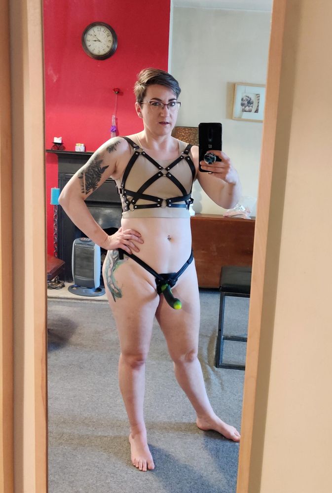 Mirror selfie of me wearing only a pale tan binder, a leather chest harness, a leather strap-on harness, and a curved strap-on cock, standing with one hand on my hip and showing off my tattoos and muscular physique.