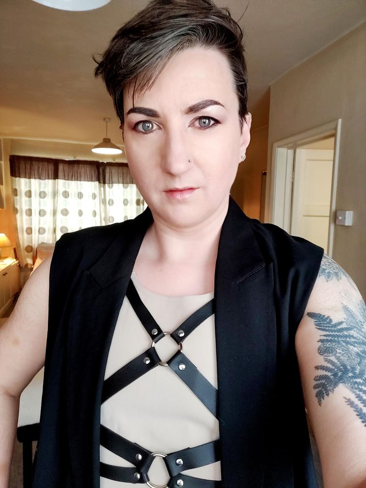 Selfie of me, a white non-binary person with short hair and tattoos, wearing a binder and a sleeveless black jacket