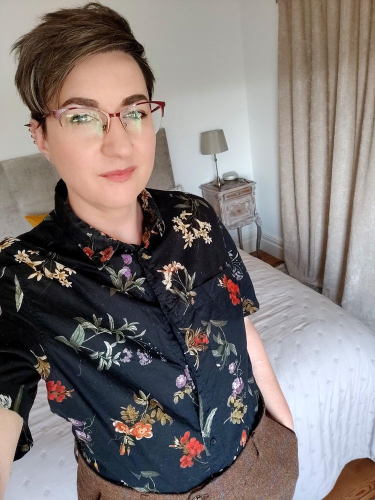 Selfie of me, a white non-binary person with short brown hair and glasses, wearing tweed trousers and a floral black short sleeved shirt