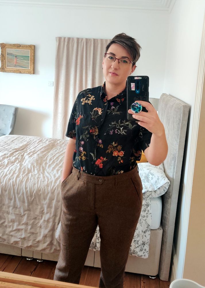 Mirror selfie of me, a white non-binary person with short brown hair and glasses, wearing tweed trousers and a floral black short sleeved shirt