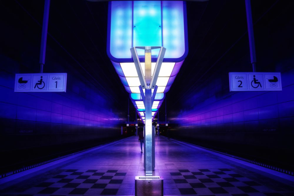 This image captures the futuristic interior of the Hafencity Universität U-Bahn station in Hamburg, Germany. The platform is illuminated by striking blue and purple lighting, creating a surreal, almost sci-fi atmosphere. A central metallic structure supports a series of glowing rectangular light panels that extend along the ceiling, adding symmetry and depth to the composition.

The walls reflect the ambient lighting, further enhancing the station’s modern aesthetic. On either side of the platform, signs indicate Gleis 1 and Gleis 2, with accessibility symbols for wheelchairs and strollers. A checkerboard pattern on the floor adds an extra layer of visual interest. In the distance, a few passengers can be seen walking, their silhouettes blending into the vibrant lighting.