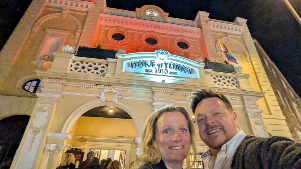 Julie and Remy pose in front of a lit up front of cinema. A yellow-ish building with intricate details around the arches, and a sign reading "Duke of York's, established 1910"