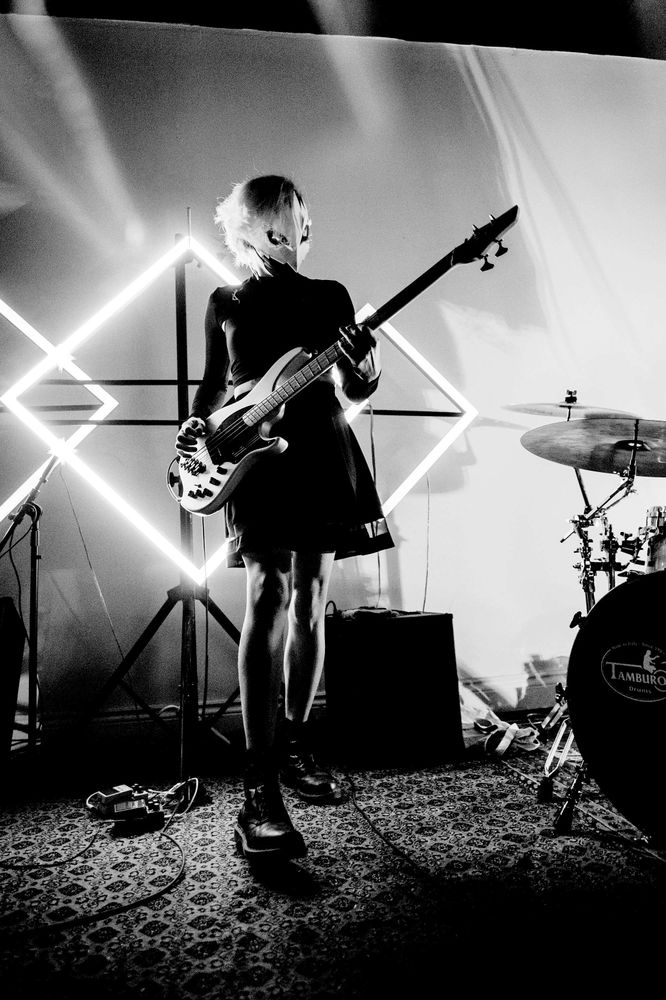 She's standing in front of diamond-shaped LED lights on a stage, playing bass guitar. Photo by Alex Carre