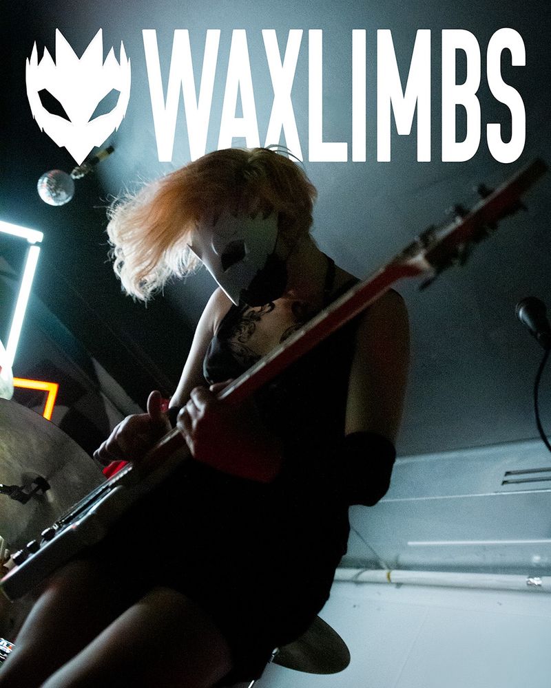 A girl in a black dress playing bass on a stage. She is wearing a white birdlike mask and is lit from behind.