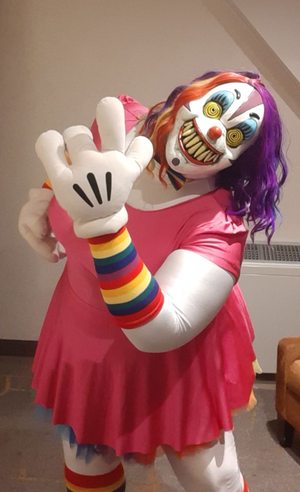 Clown doll girl cosplay in pink dress and 3 finger toon gloves. Also has rainbow hair, socks and legwarmers. Mask has sharp teeth, yellow swirly eyes and evil grin.