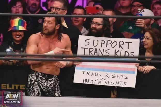 Kenny omega in front of a support trans kids sign