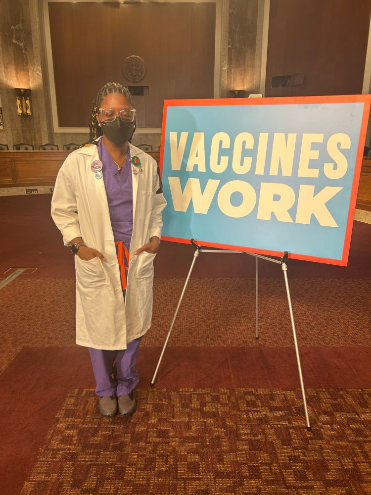 Picture of Lawren, a Black woman in purple scrubs and long white coat wearing a trans flag pin, watermelon pin, "end gun violence" pin and "bans off our bodies" pin. She stands next to a large sign that reads "vaccines work".