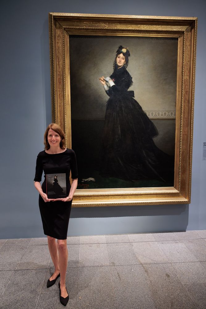 Redheaded woman in black dress stands holding book with the same cover as the painting behind her, which depicts a woman in a 19th c. black dress staring out.