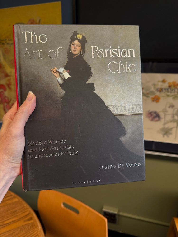 Cover of The Art of Parisian Chic: Modern Women and Modern Artists in Impressionist Paris with a woman standing, staring out and removing one glove. She wears a black dress and has a prominent tea rose in her hat. Vague office setting behind.
