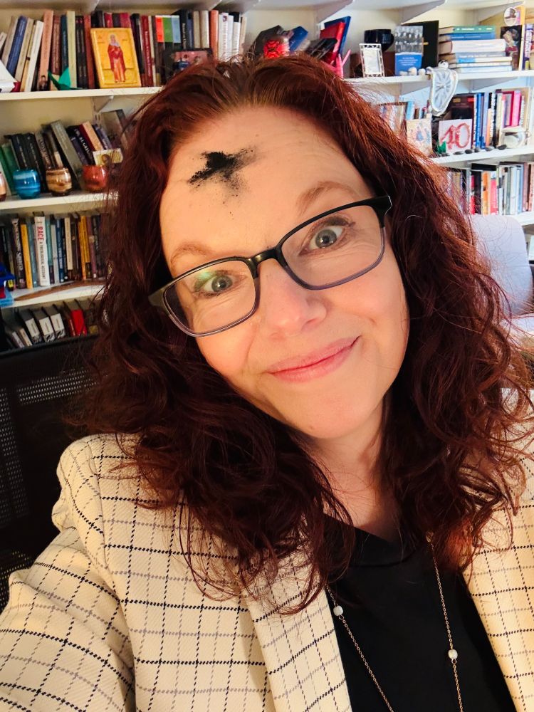 Selfie of a red--haired white woman with ashes in the shape of the cross on her forehead in an academic office lined with books 