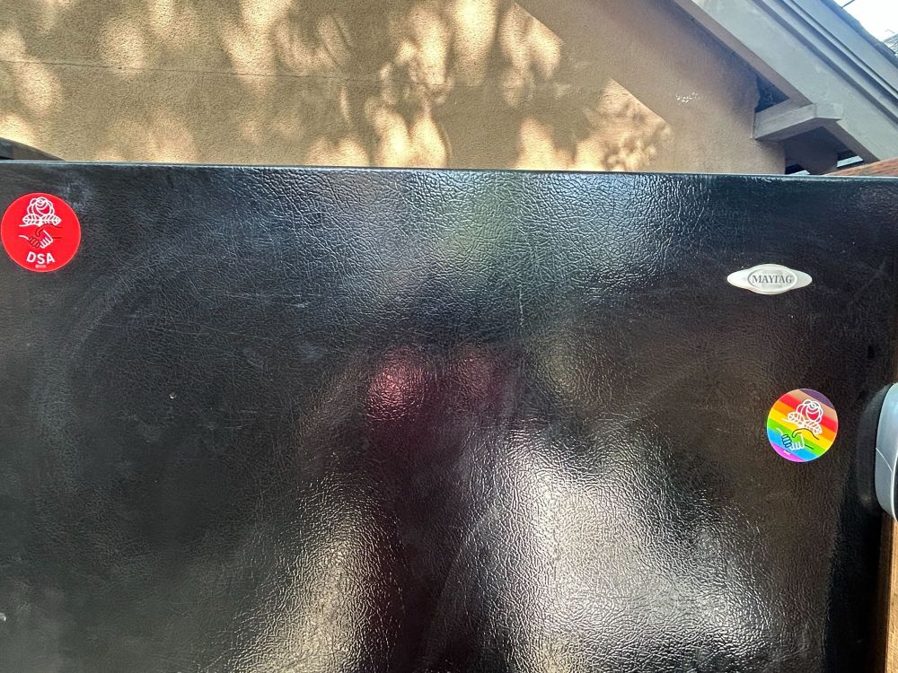 Detail of the black fridge, adorned with two stickers. One is the standard red, and the other has LGBTQAI+ rainbow colors