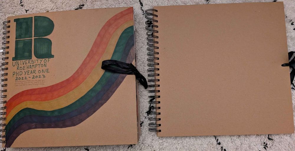 A photograph of two scrapbooks. The one on the left features a rainbow sprawling across the bottom left corner to the top right corner. In the top left corner is the logo for my University, and text that reads PhD Year One.