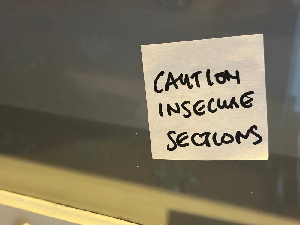 A handwritten sign, with black ink on white paper and stuck on a window, which says “Caution insecure sections”