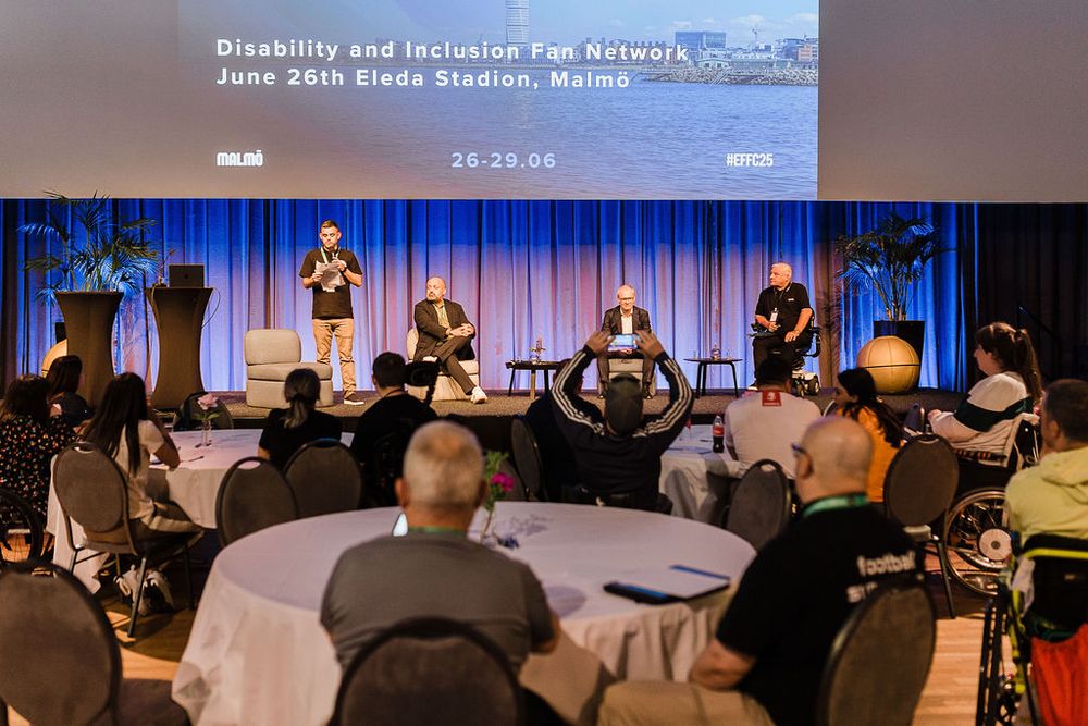 A panel discussion in progress on stage at the Disability and Inclusion Fan Network event in Malmö. Four speakers, including one in a wheelchair, are seated on stage with a large screen behind them displaying event details. Audience members, some in wheelchairs, are seated at round tables, listening attentively or taking photos.