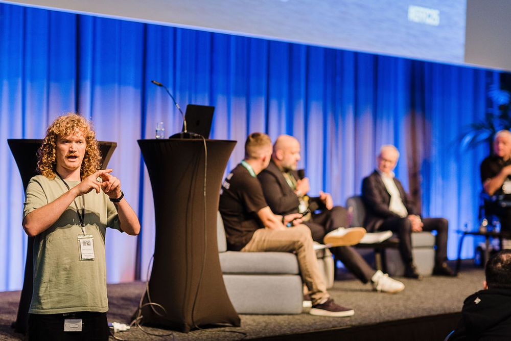 A man is centre-left gesturing sign language. He wears a light green t-shirt and a lanyard. In the background to the right, four men are seated on a stage against a bright blue curtain, engaged in a panel discussion.